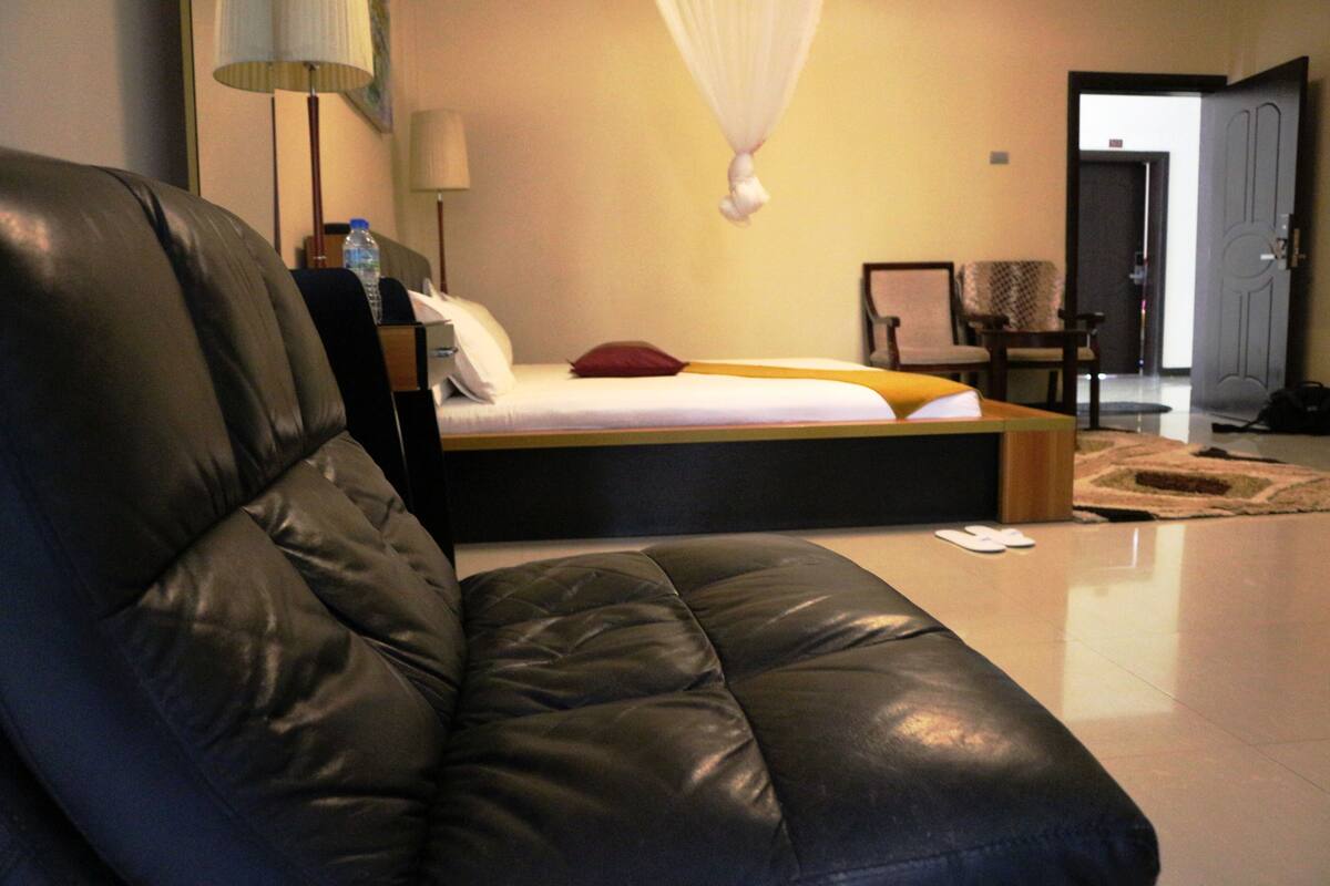 A modern hotel room with a black leather sofa, a bed with white bedding, a bedside table with a lamp, and a door leading to another room.