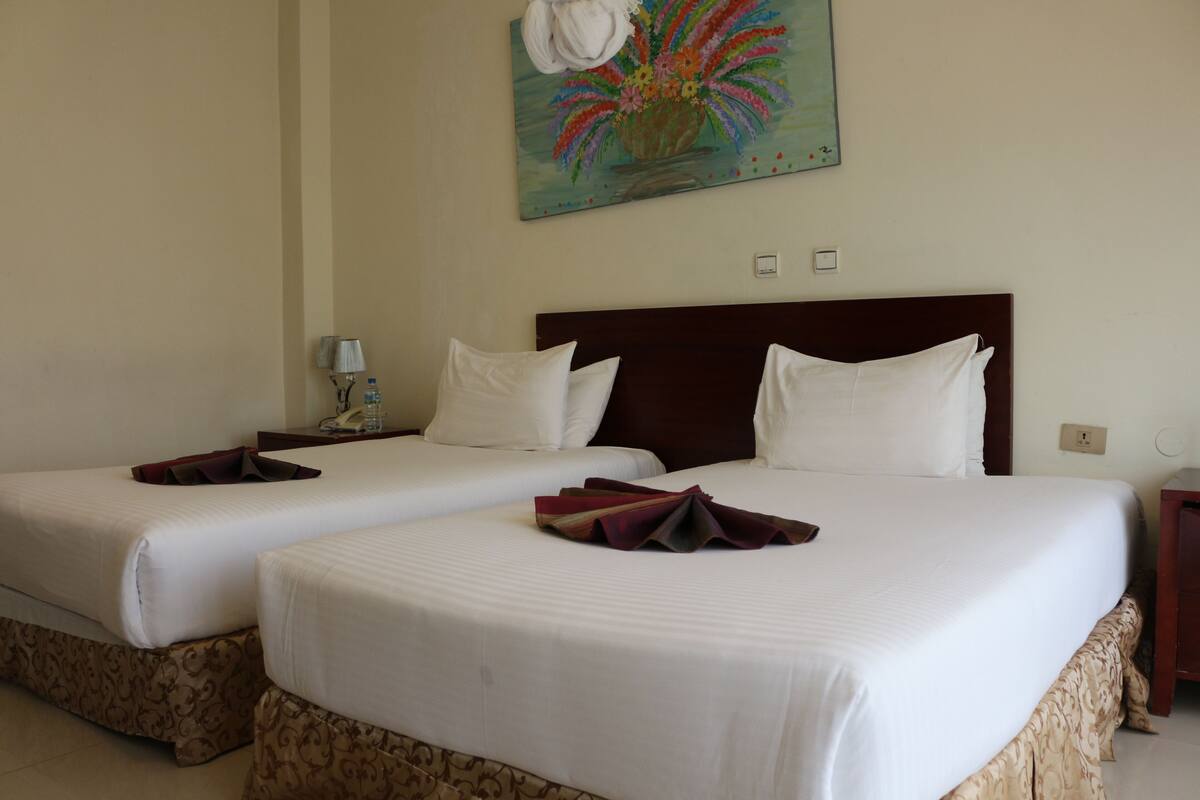 A hotel room with two beds, each with white linens and a dark headboard. There is a painting of flowers on the wall above the beds.