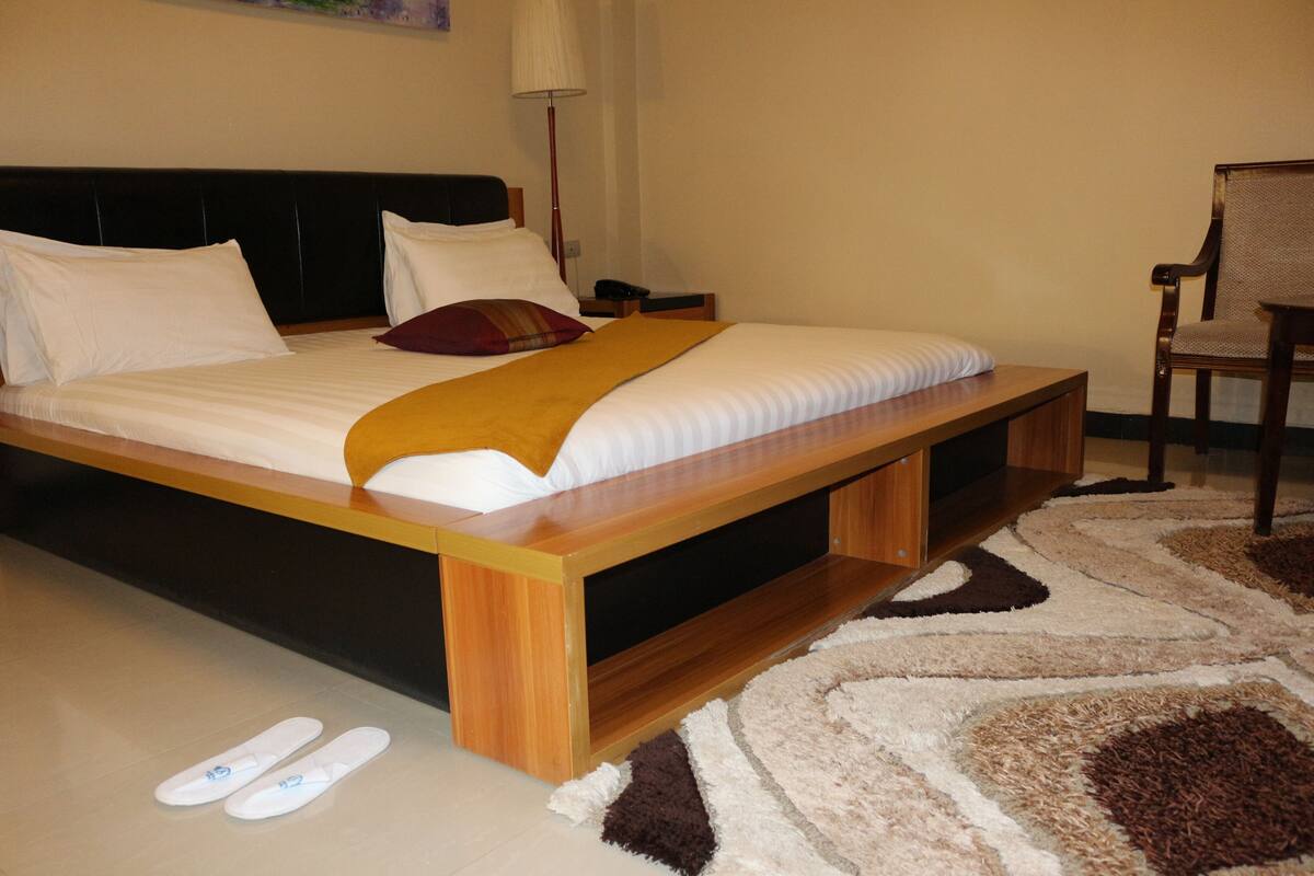 A bed with a wooden frame, white bedding, and a brown throw blanket. There is a chair and a lamp in the room.