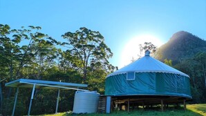 A yurt with a covered entrance, a white structure with a roof, and a water tank in a grassy area with trees and a hill in the background. - NOOSA ECO RETREAT (Pomona)