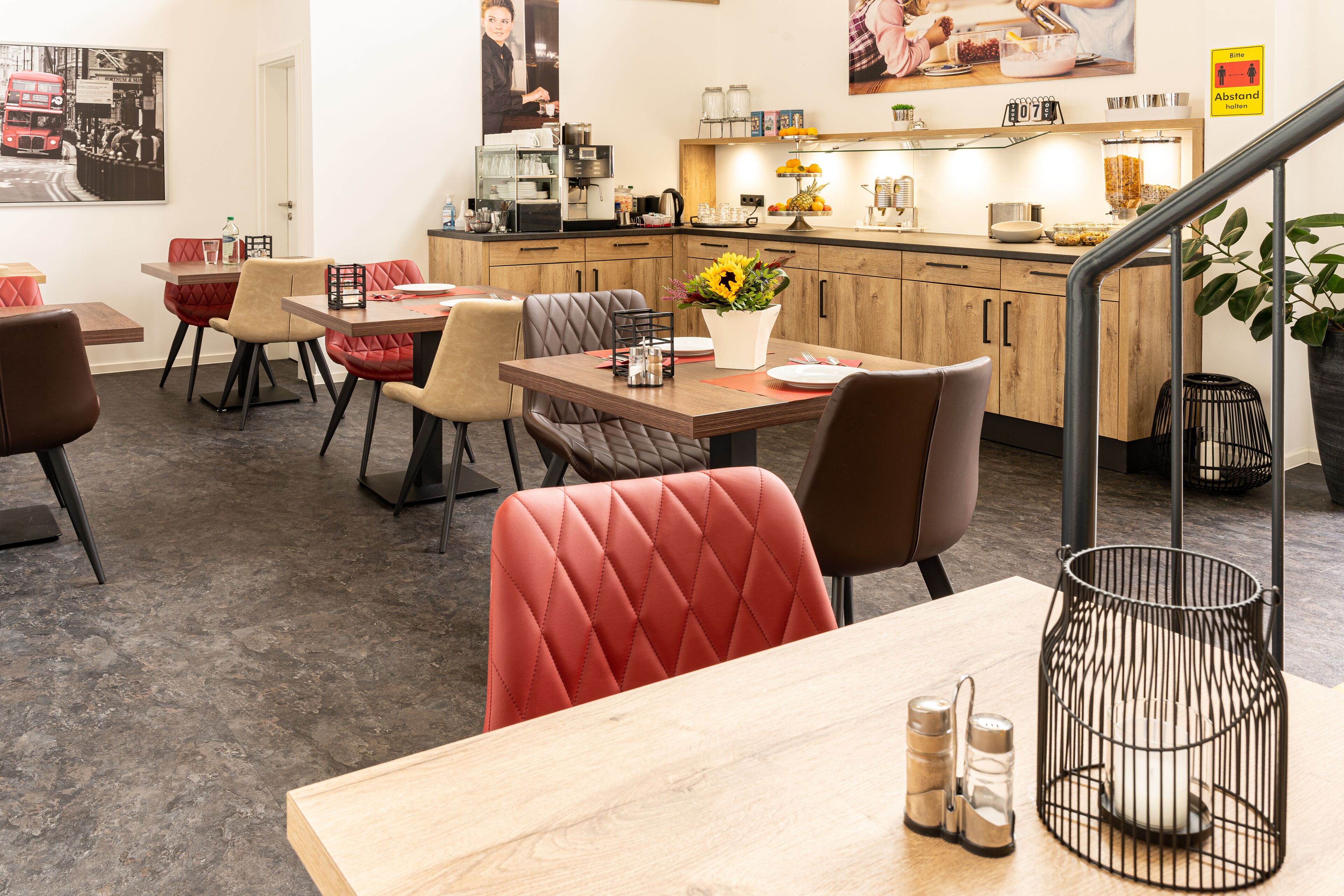 A modern dining area with wooden tables and red cushioned chairs, a coffee station, and a kitchen in the background.