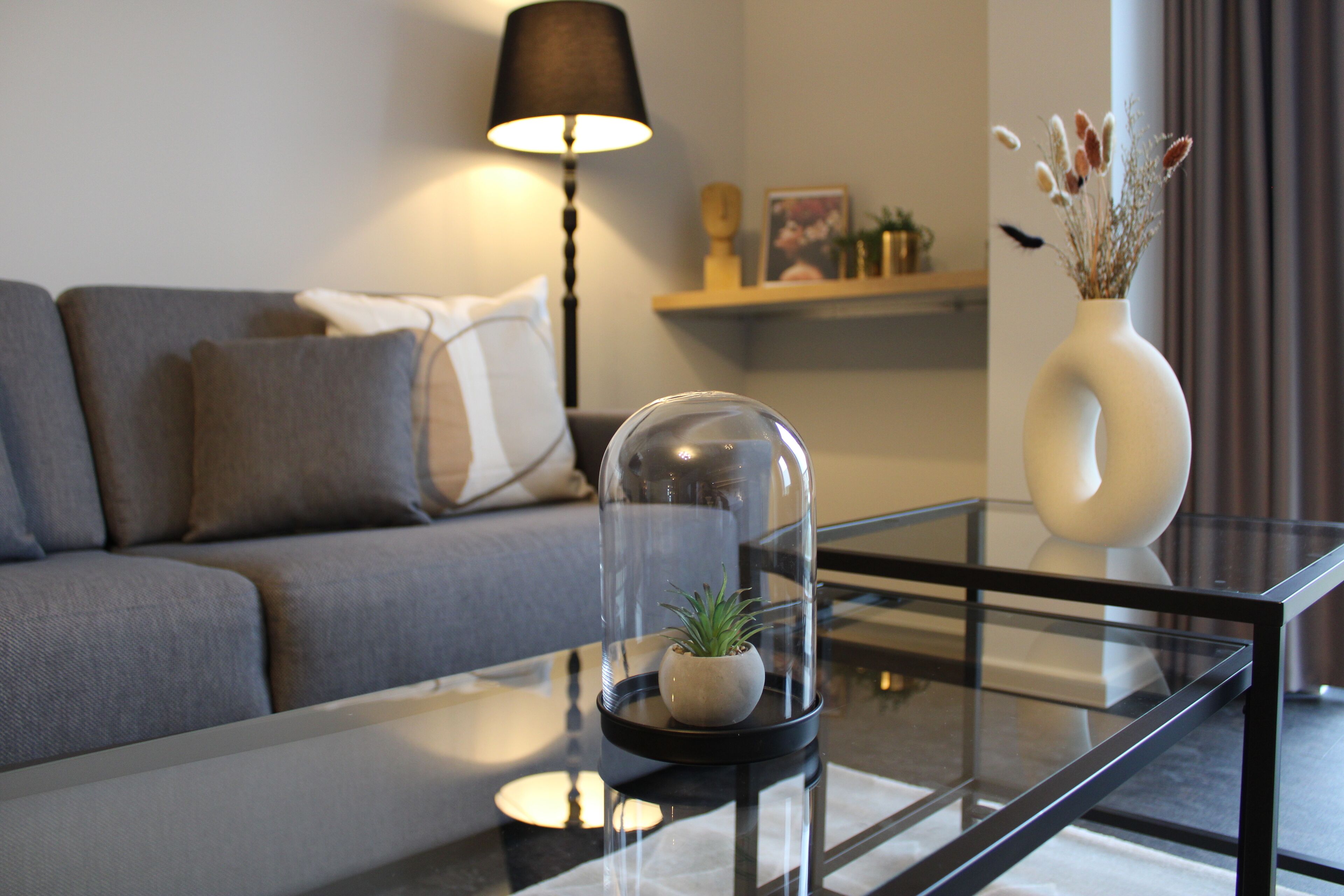 Una sala de estar moderna con una mesa de centro de vidrio, un sofá, una lámpara y un jarrón con flores secas.