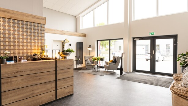 A modern reception area with a wooden reception desk, two seating areas with chairs and tables, and large windows providing natural light.