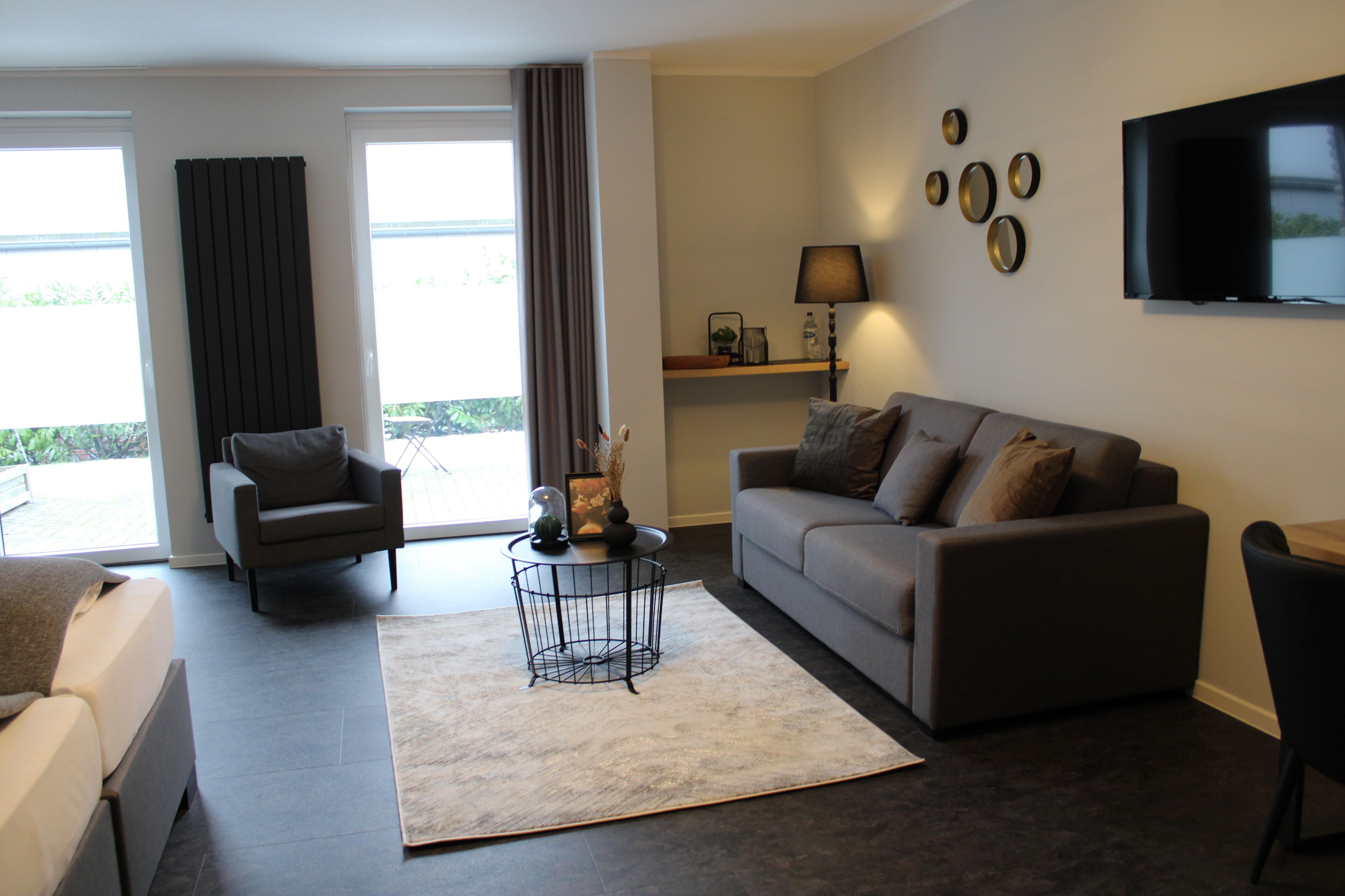 A modern living room with a grey sofa, a black armchair, and a coffee table. There is a flat-screen TV mounted on the wall and a window with curtains.