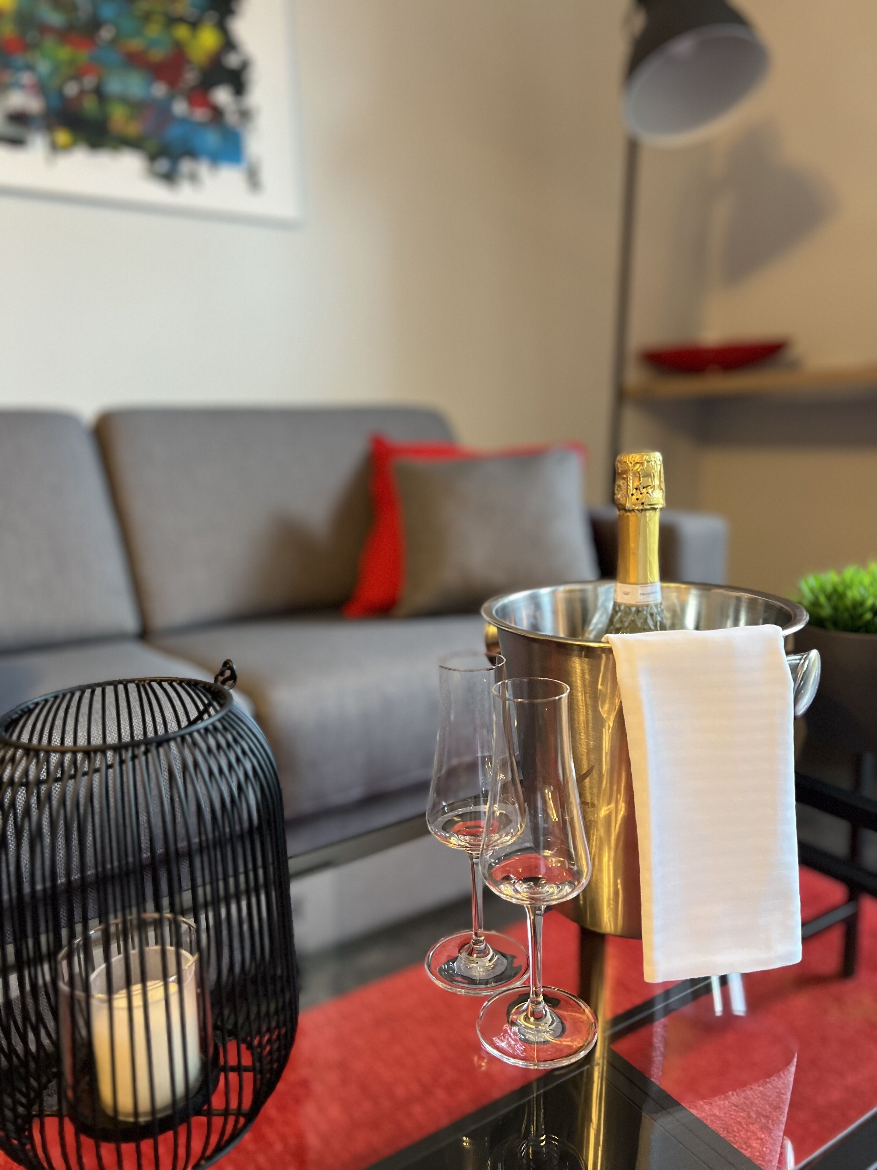 A modern living room with a sofa, a red rug, a glass table with a bottle of champagne, two wine glasses, and a candle in a black lantern.