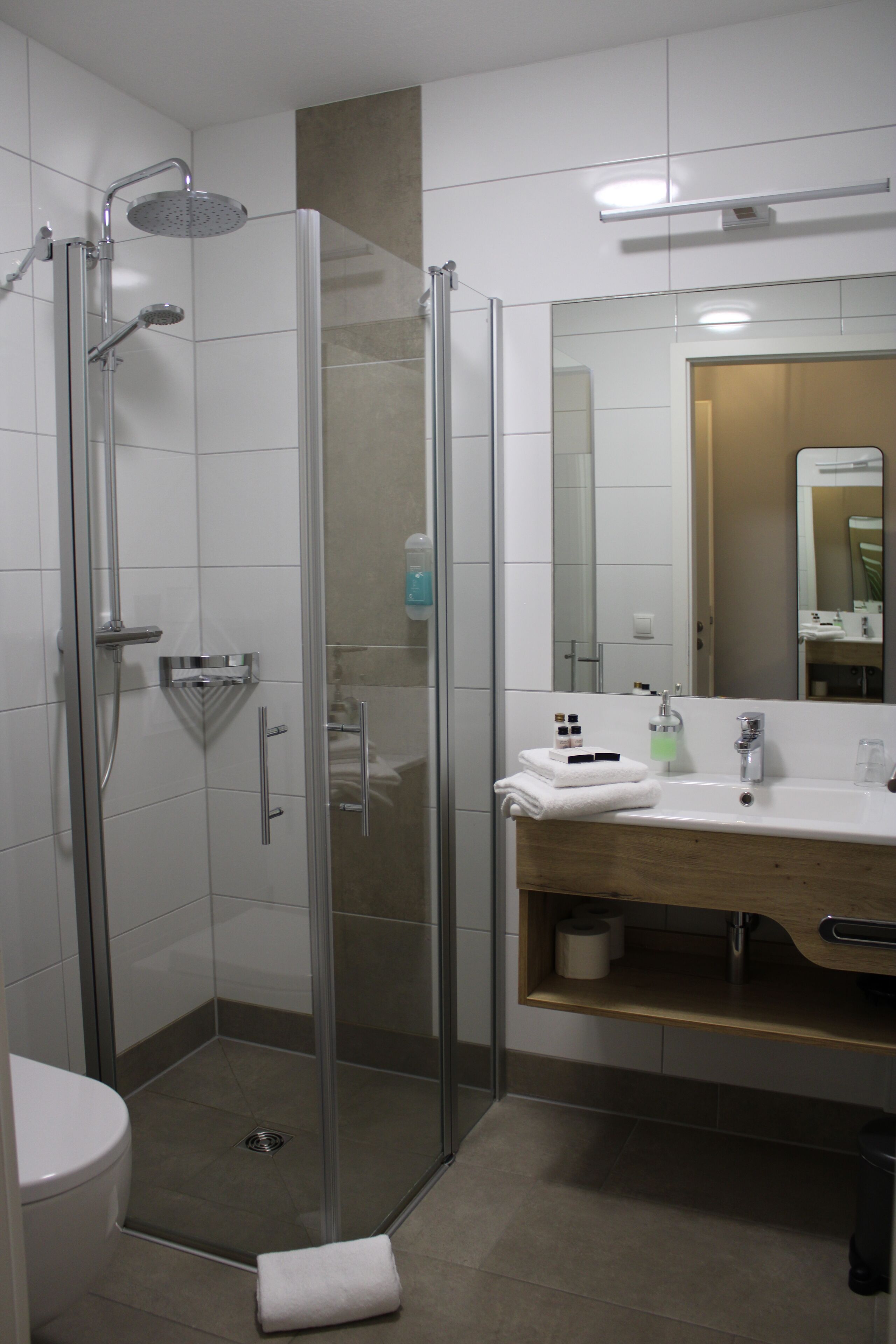 Un baño moderno con ducha envuelta en vidrio, un lavabo blanco con tocador de madera y un espejo grande sobre el lavabo.