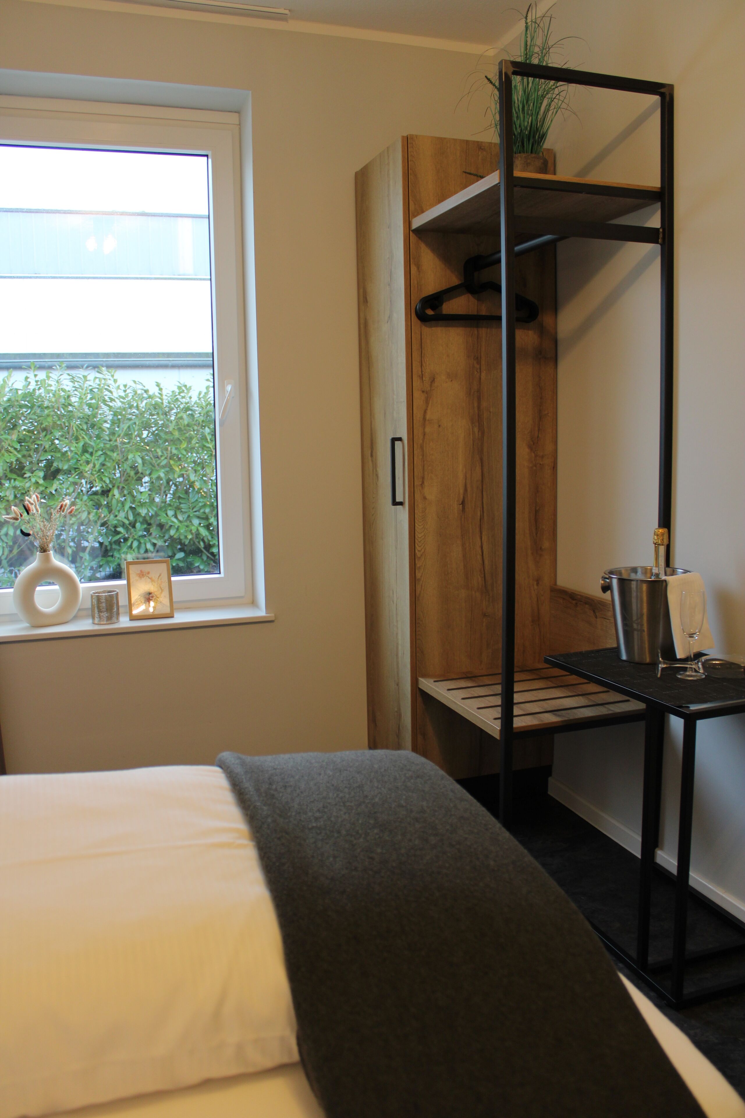A bedroom with a bed, a window with a view of greenery, a wooden cabinet, and a metal shelf with a plant.