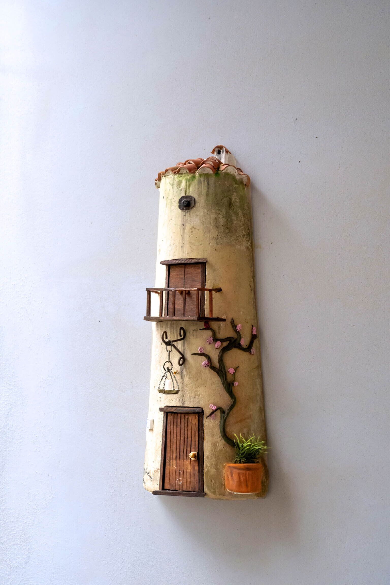 Una pieza decorativa montada en la pared con un diseño rústico, que presenta un pequeño ventanal, una puerta y una planta en maceta.