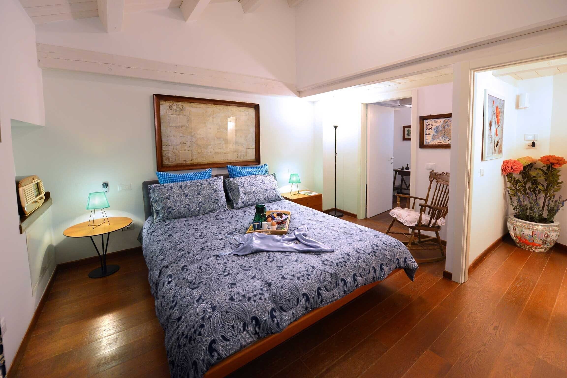 Quarto com cama grande, mesa de cabeceira com abajur, piso de madeira e porta que dá para outro cômodo.