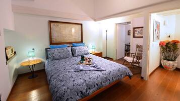 Quarto com cama grande, mesa de cabeceira com abajur, piso de madeira e porta que dá para outro cômodo.