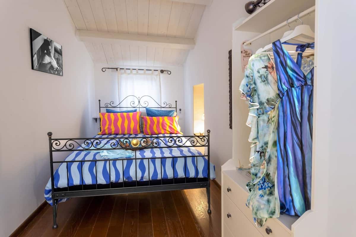 A bedroom with a metal bed frame, striped bedding, a window with curtains, and a clothing rack with a floral dress.