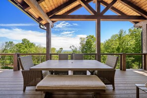 Una terraza de madera al aire libre con una mesa y sillas, rodeada por barandilla de madera, con vistas a un paisaje verde.