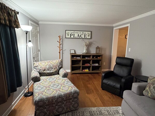 Una sala de estar con un reposapiés estampado, un sillón de cuero negro, una estantería y un cartel en la pared que dice 'Welcome to Our PLACE'.