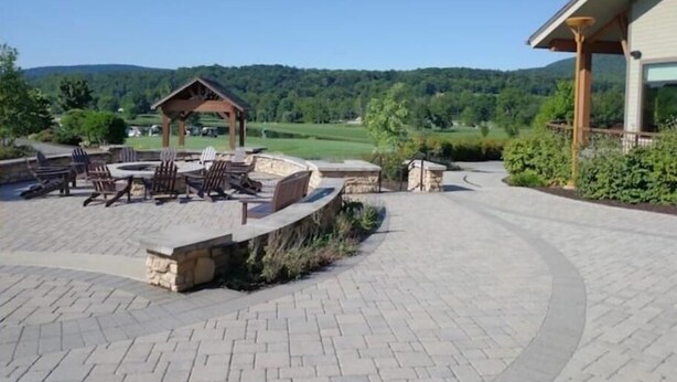 Un patio con mobiliario exterior y una chimenea de piedra, rodeado por un camino pavimentado y vistas a un paisaje verde.