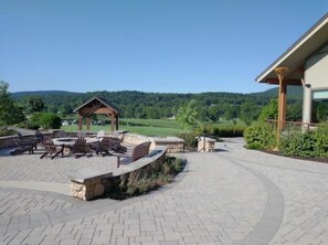 Eine Terrasse mit Gartenmöbeln und einem Steinkamin, umgeben von einem gepflasterten Weg und Blick auf eine grüne Landschaft.