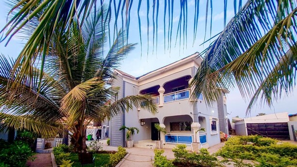 A two-story building with a balcony, surrounded by greenery and palm trees.