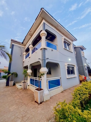 A two-story building with a balcony, a covered patio area, and a small garden with potted plants. - Sun Rise Apt #5 The Retreat Apartments (Brusubi)