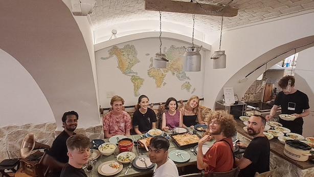 Un groupe de personnes est réuni autour d’une table dans une pièce où l’on peut voir une grande carte accrochée au mur, ainsi que des meubles en bois et des pots suspendus.