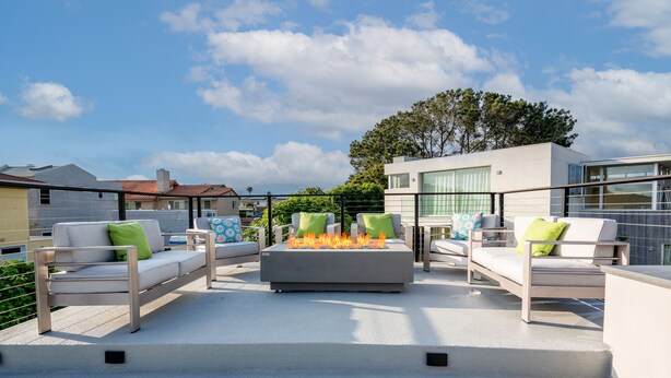 An outdoor patio with a modern fire feature, two sofas with cushions, and a view of neighboring buildings. - Ocean View Rooftop Deck + Walk to Beach (Cardiff By The Sea)