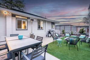 Une maison moderne dotée d’un patio comprenant une table et des chaises, un barbecue et une pelouse bien entretenue.