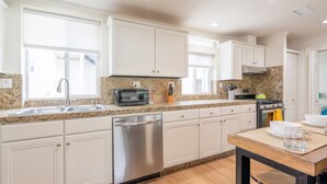 A modern kitchen with white cabinets, granite countertops, stainless steel appliances, and a dining area with a wooden table. - Ocean View Rooftop Deck + Walk to Beach (Cardiff By The Sea)