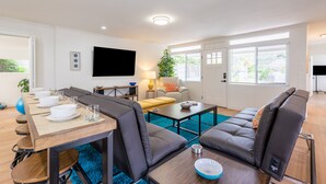 A modern living room with a dining area, a sofa set, a flat-screen TV, and a large window with blinds. - Ocean View Rooftop Deck + Walk to Beach (Cardiff By The Sea)