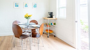 A dining area with a white brick wall, a wooden table with chairs, a coffee maker, and a dish rack with colorful dishes. - Ocean View Rooftop Deck + Walk to Beach (Cardiff By The Sea)