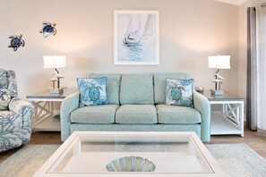 A living room with a light blue sofa, two matching pillows, a framed seascape painting, a glass coffee table, and two side tables with lamps. - Oceanfront 2 BR Condo with Shared Pool! Sea Shadow 406  (Surfside Beach)