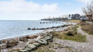 A coastal path with a rocky shoreline, a wooden pier, and a row of houses in the background.