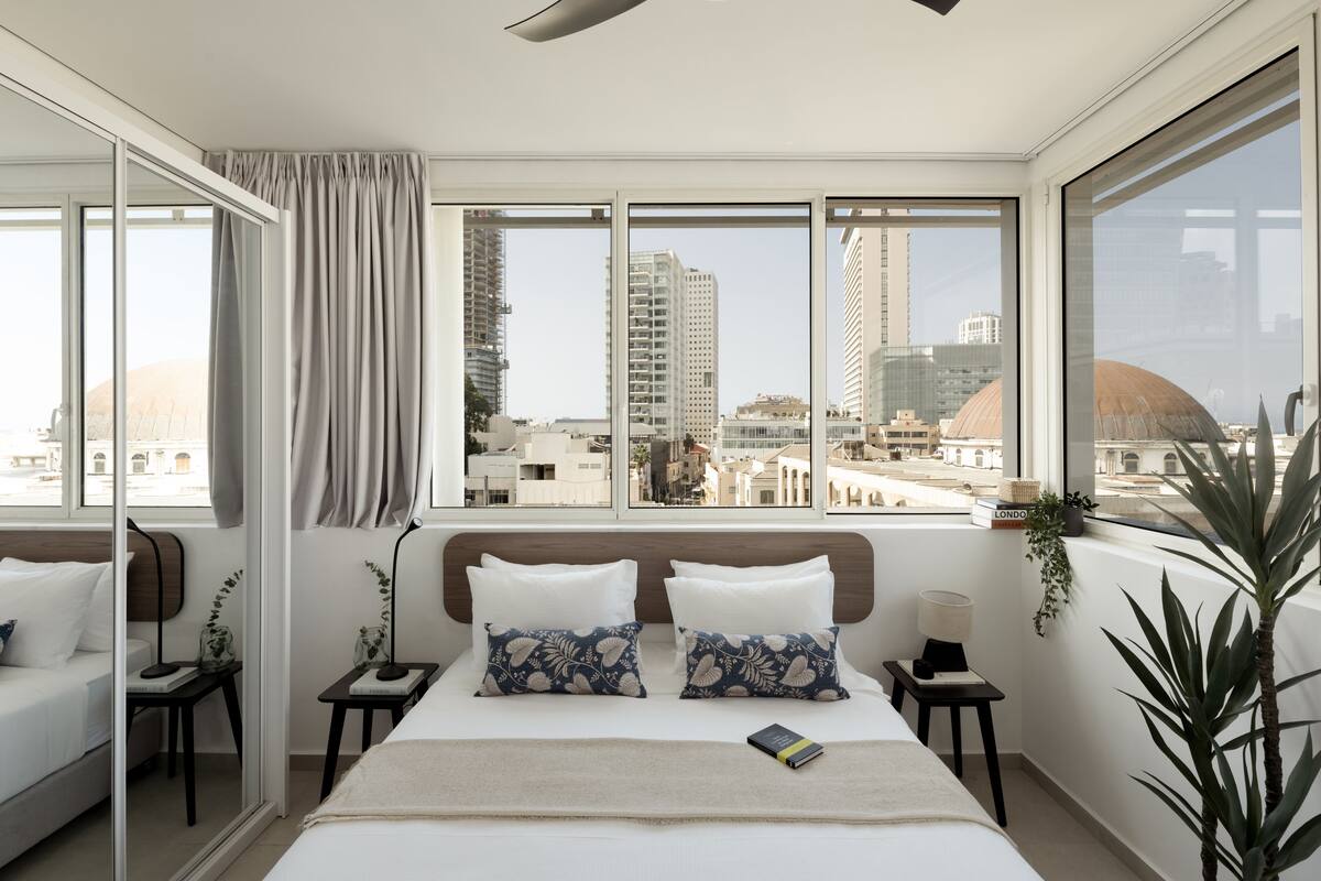 Un hotel moderno con una cama grande, dos mesitas de noche, un espejo y vistas de la ciudad a través de ventanas amplias.