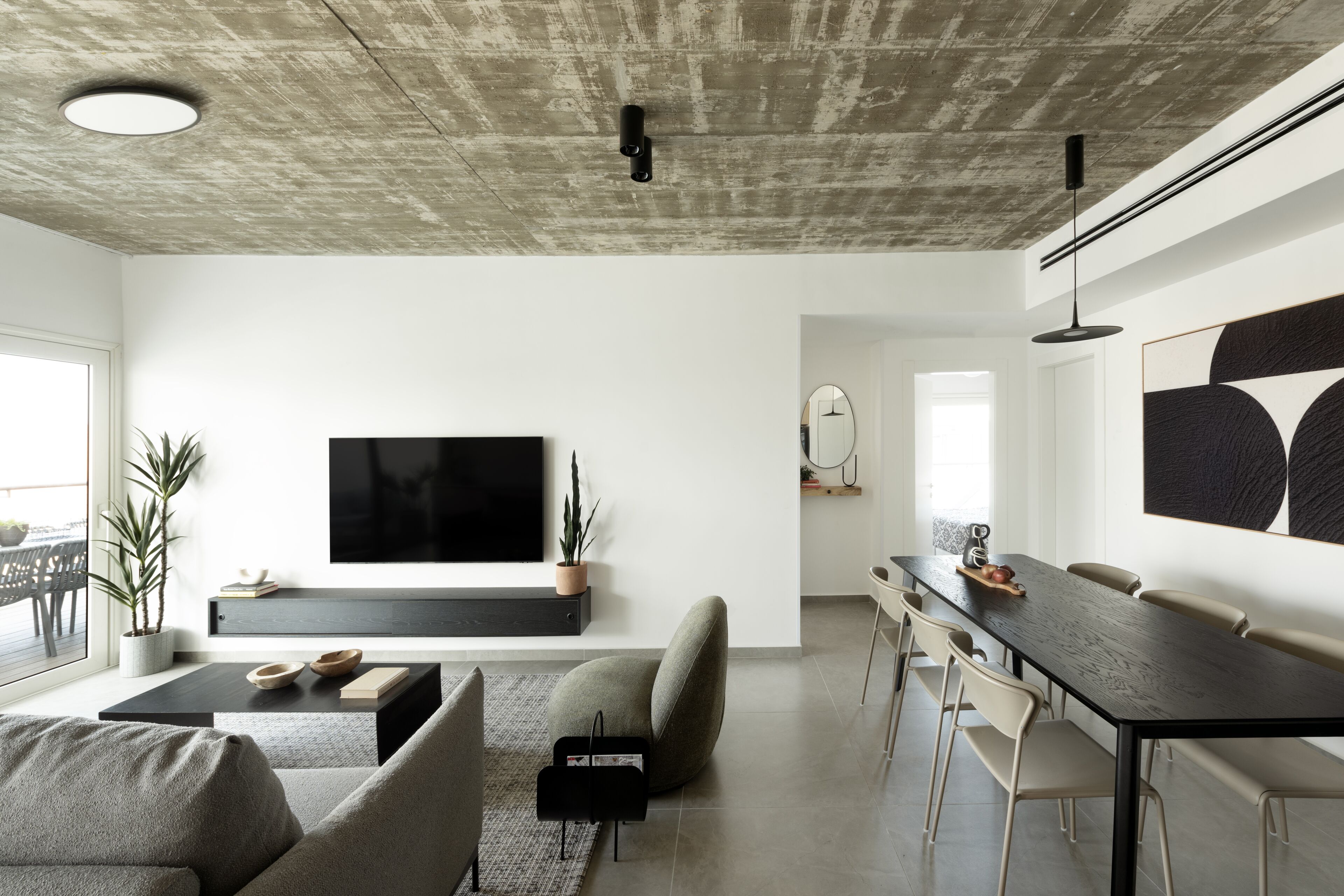 Ein modernes Wohnzimmer mit einem großen Flachbildfernseher, einem Essbereich mit langem Tisch und Stühlen sowie einem minimalistischen Design mit Betondecke und weißen Wänden.