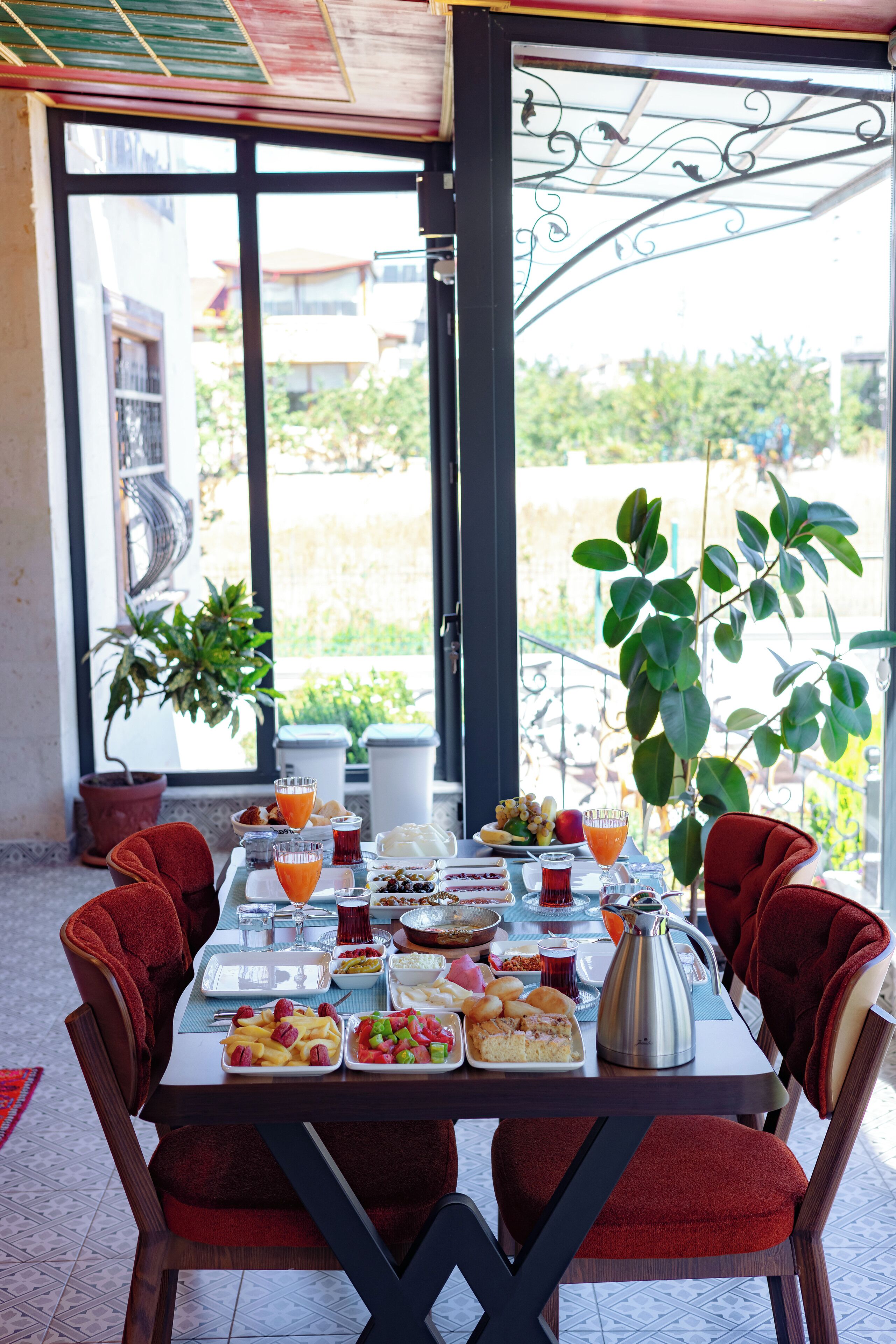 A dining table set with various dishes and drinks, surrounded by chairs, in a room with large windows and potted plants.