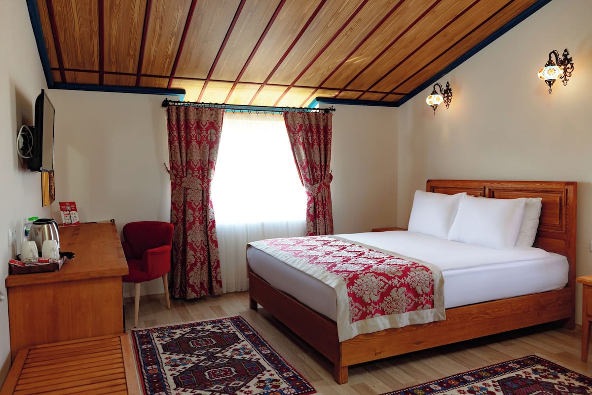 Een slaapkamer met een houten bed, rode gordijnen, een rode stoel, een houten bureau en een kamerkleed met een patroon.