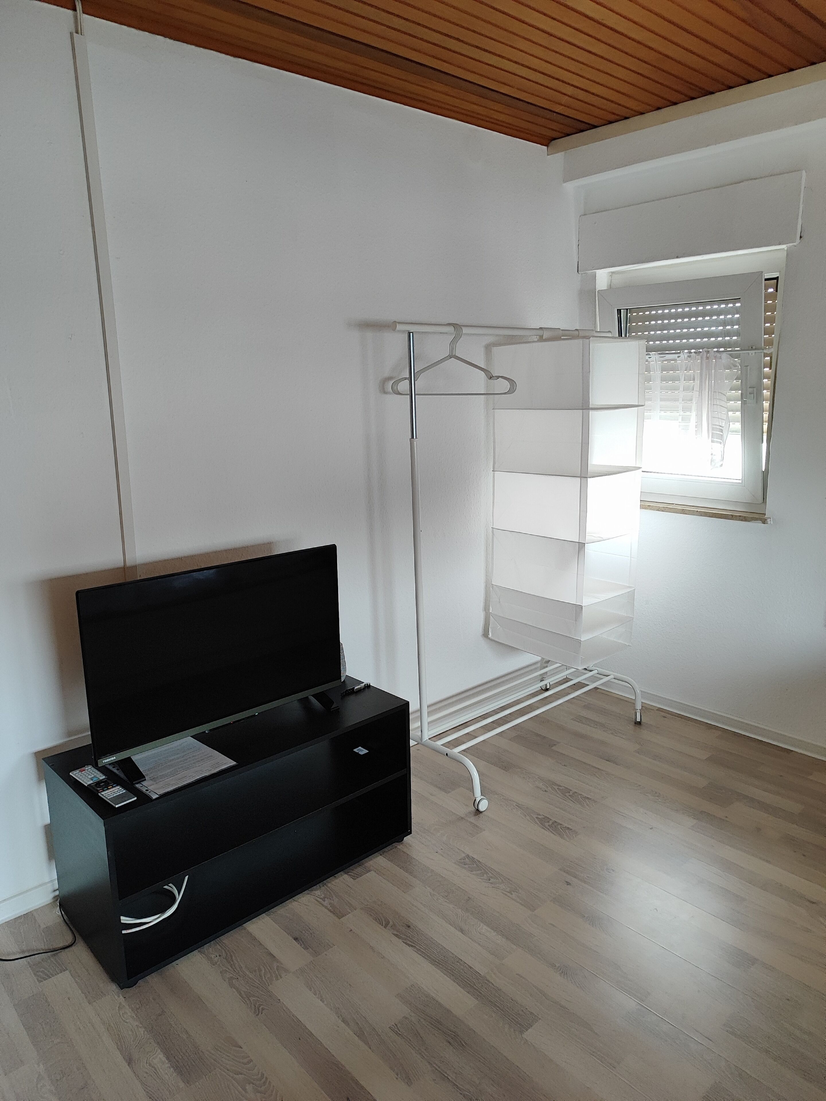Une pièce avec un plafond en bois, un meuble de rangement noir pour appareils électroniques, une armoire blanche avec une tringle, et une fenêtre avec des stores.
