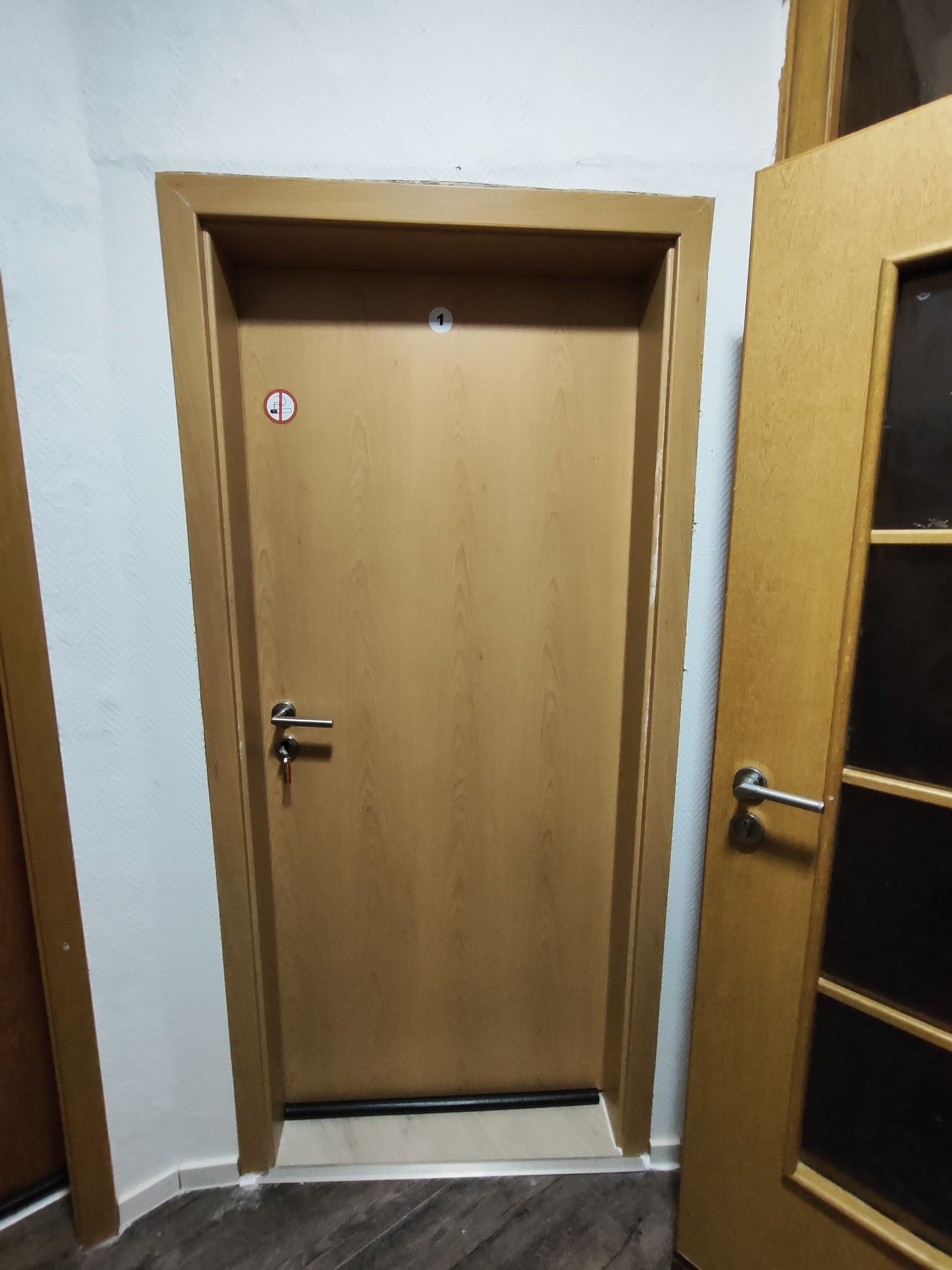 Una puerta de madera cerrada con manija metálica y cerradura, ubicada en una habitación con paredes blancas y suelo oscuro.