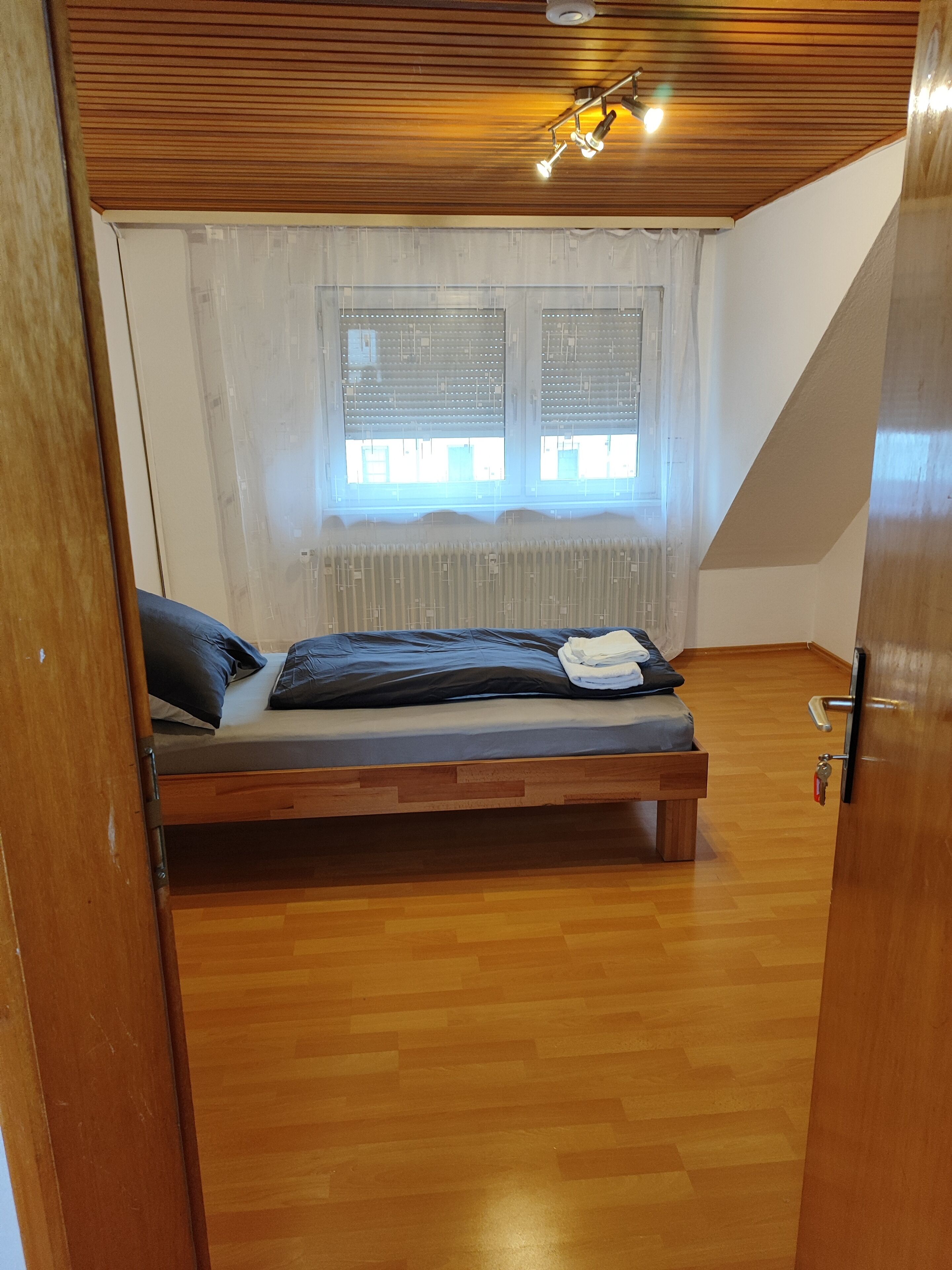 Una stanza con pavimento in legno, un letto con materasso e cuscini, una finestra con persiane e un soffitto con faretti.