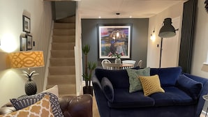 Una sala de estar con un sofá azul oscuro, un sofá de cuero marrón y piso de madera. Hay una escalera que lleva a otra habitación, una mesa de comedor con sillas y un gran ventanal con vistas.