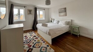 Ein Schlafzimmer mit einem Bett, einem Nachttisch, einer Lampe, einem Fenster mit Vorhängen, einem Spiegel und einem farbenfrohen Teppich.