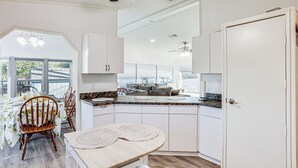 Eine moderne Küche mit weißen Schränken, ein Essbereich mit einem Holztisch und Stühlen sowie ein Blick ins Freie durch große Fenster.