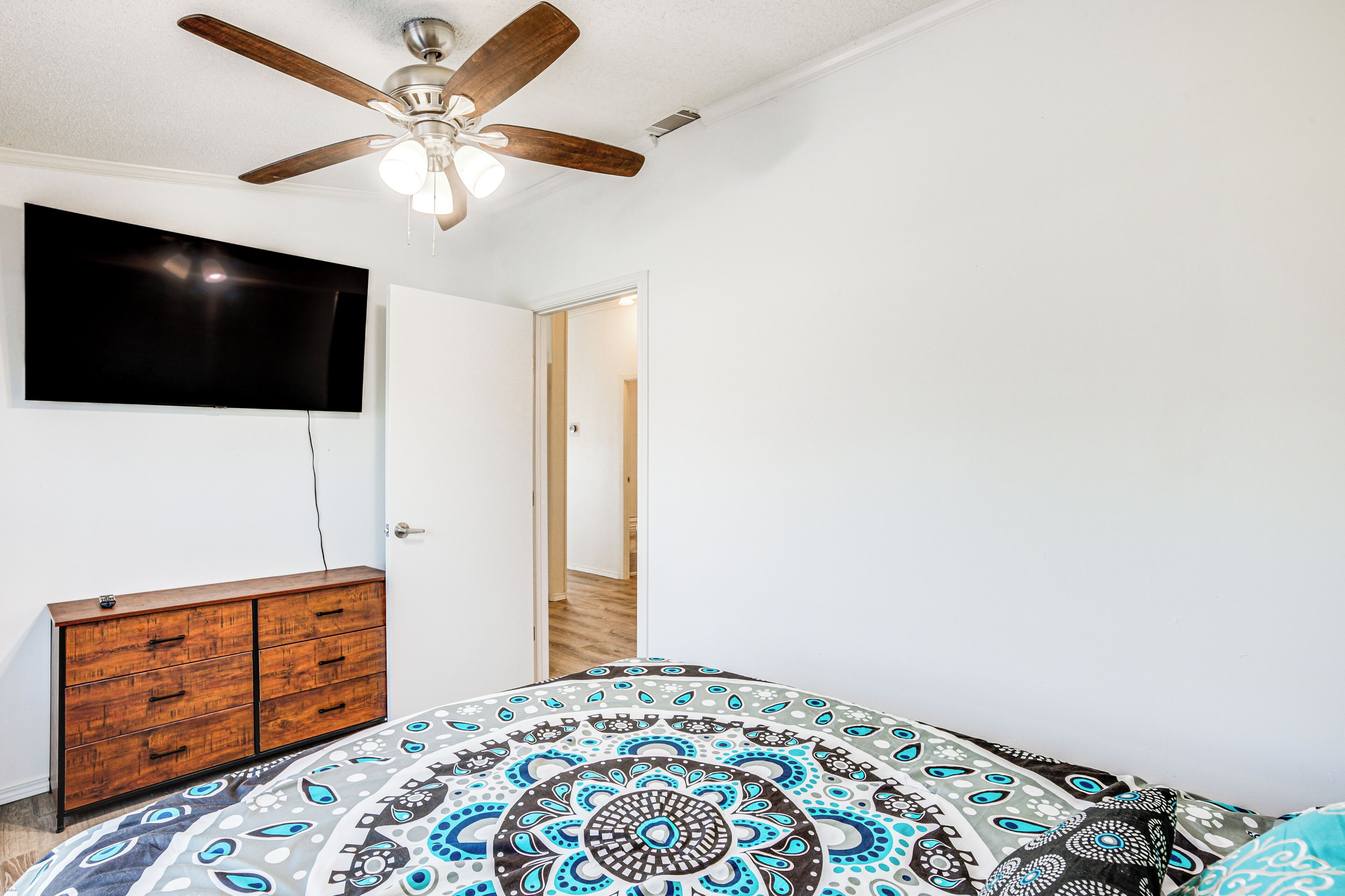 Une chambre dotée d’un ventilateur de plafond, d’un téléviseur à écran plat fixé au mur, d’une commode en bois et d’une couvre-lit à motifs.