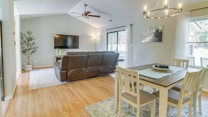A modern living room with a brown leather sofa, a wooden dining table with white chairs, a chandelier, and a flat-screen TV mounted on the wall. - Lovely Charlotte Home w/ Yard: 9 Mi to Uptown! (Charlotte)