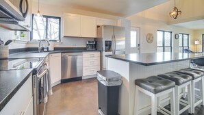 A modern kitchen with white cabinets, stainless steel appliances, a large island with bar stools, and a view of the outdoors through the windows. - Deck + Kayaks: Grand Lake of the Cherokees Home (Afton)