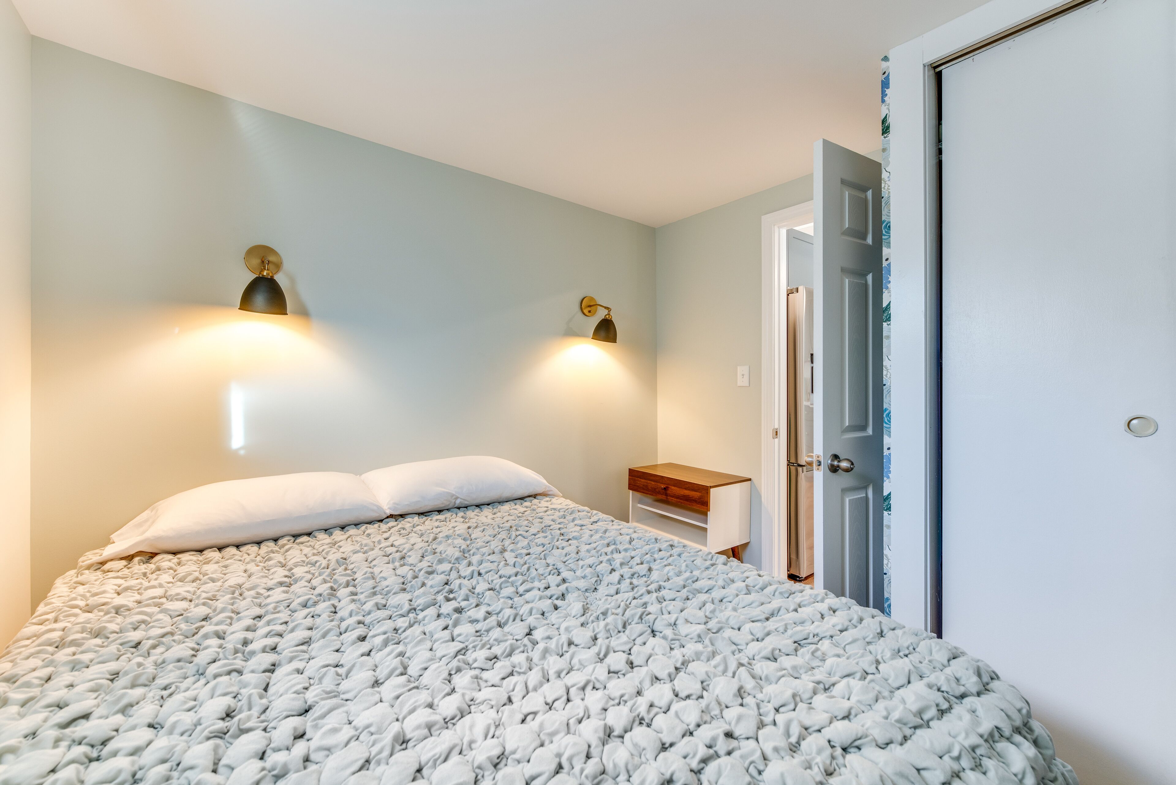 A bedroom with a large bed, two wall-mounted lights, a nightstand, and a door with a floral pattern.