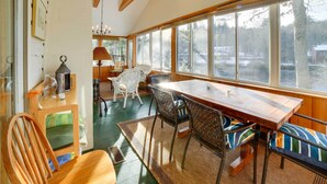 A dining area with a wooden table and chairs, large windows with a view of trees and a building, and a chandelier. - Lakefront Bradford Cottage w/ Dock, Near Skiing! (Bradford)