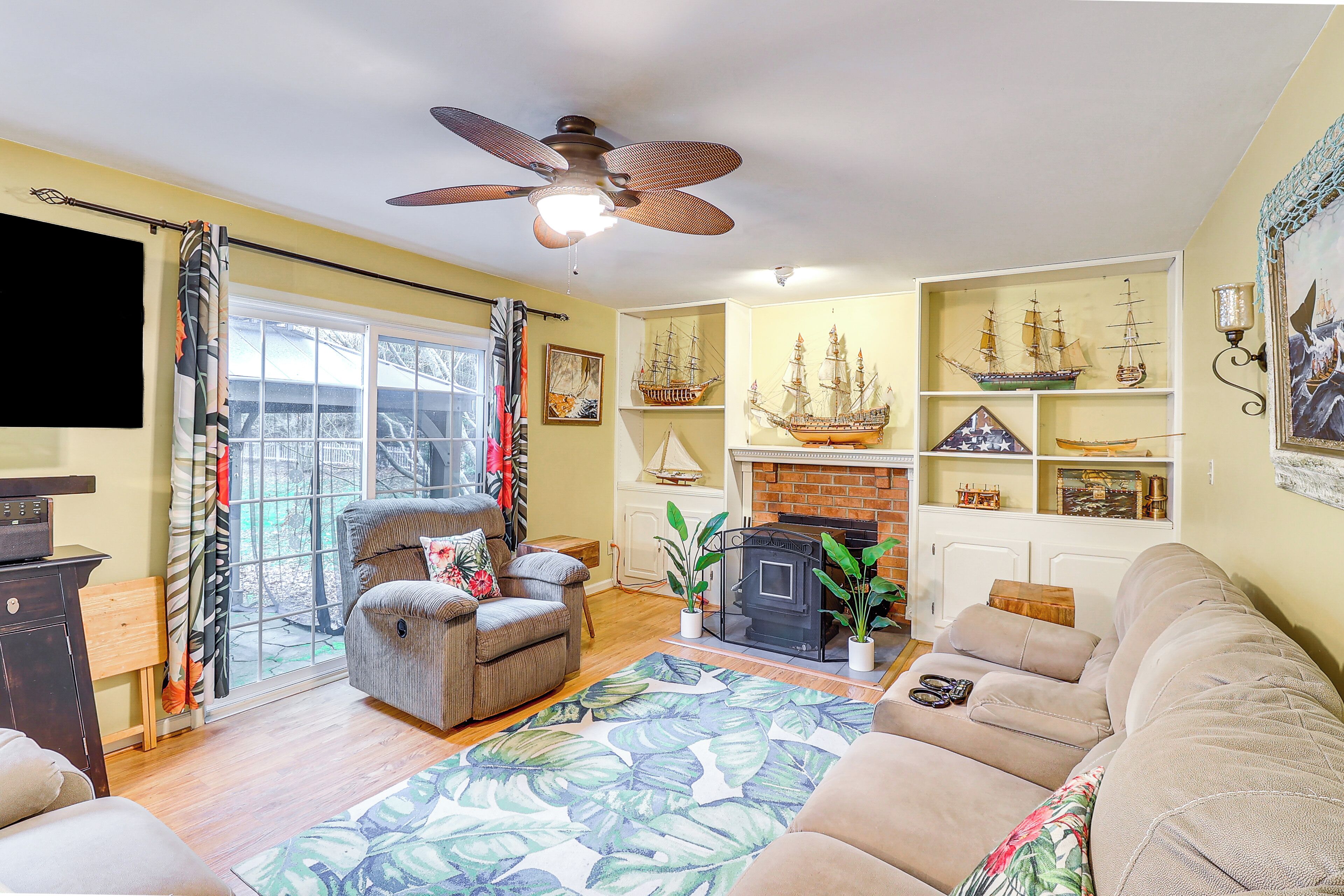 A living room with a fireplace, a ceiling fan, and a large window with curtains.