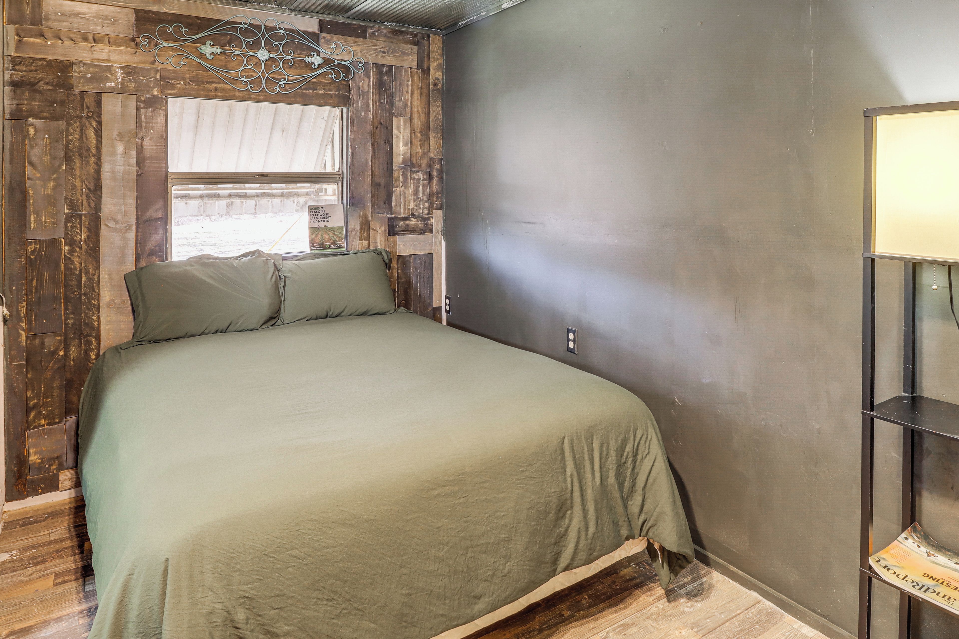 A bedroom with a wooden headboard, a bed with green bedding, a metal shelf unit, and a window with a view of the outside.