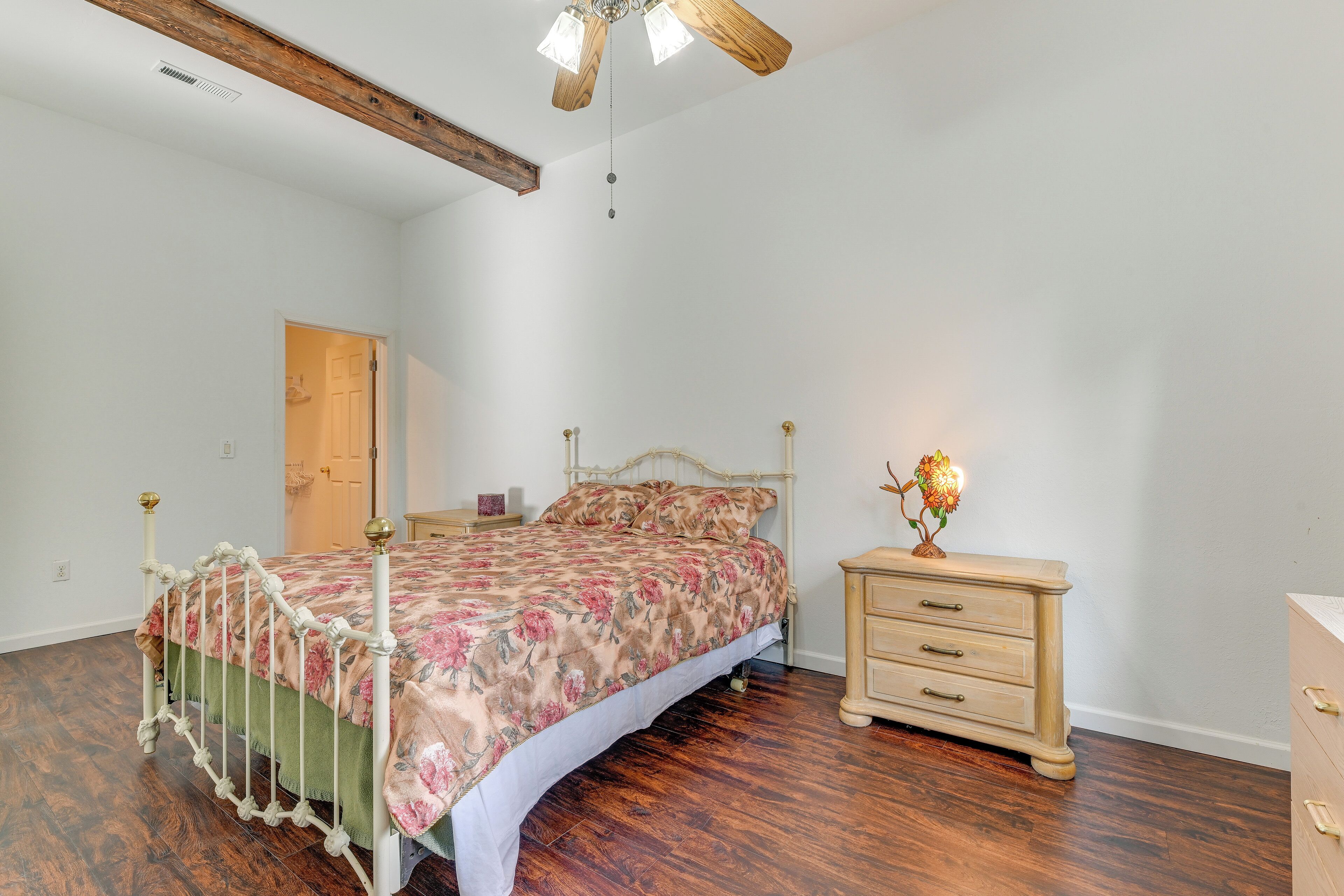 A bedroom with a wooden floor, a bed with a floral bedspread, a nightstand with a lamp, and a wooden dresser.