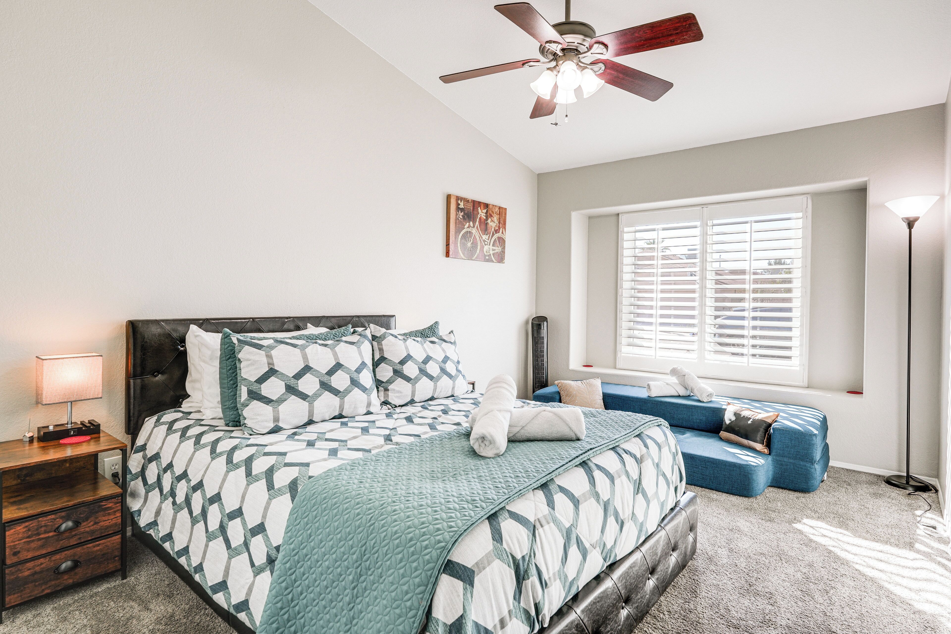 A bedroom with a bed, a nightstand, a blue sofa, a ceiling fan, and a window with blinds.
