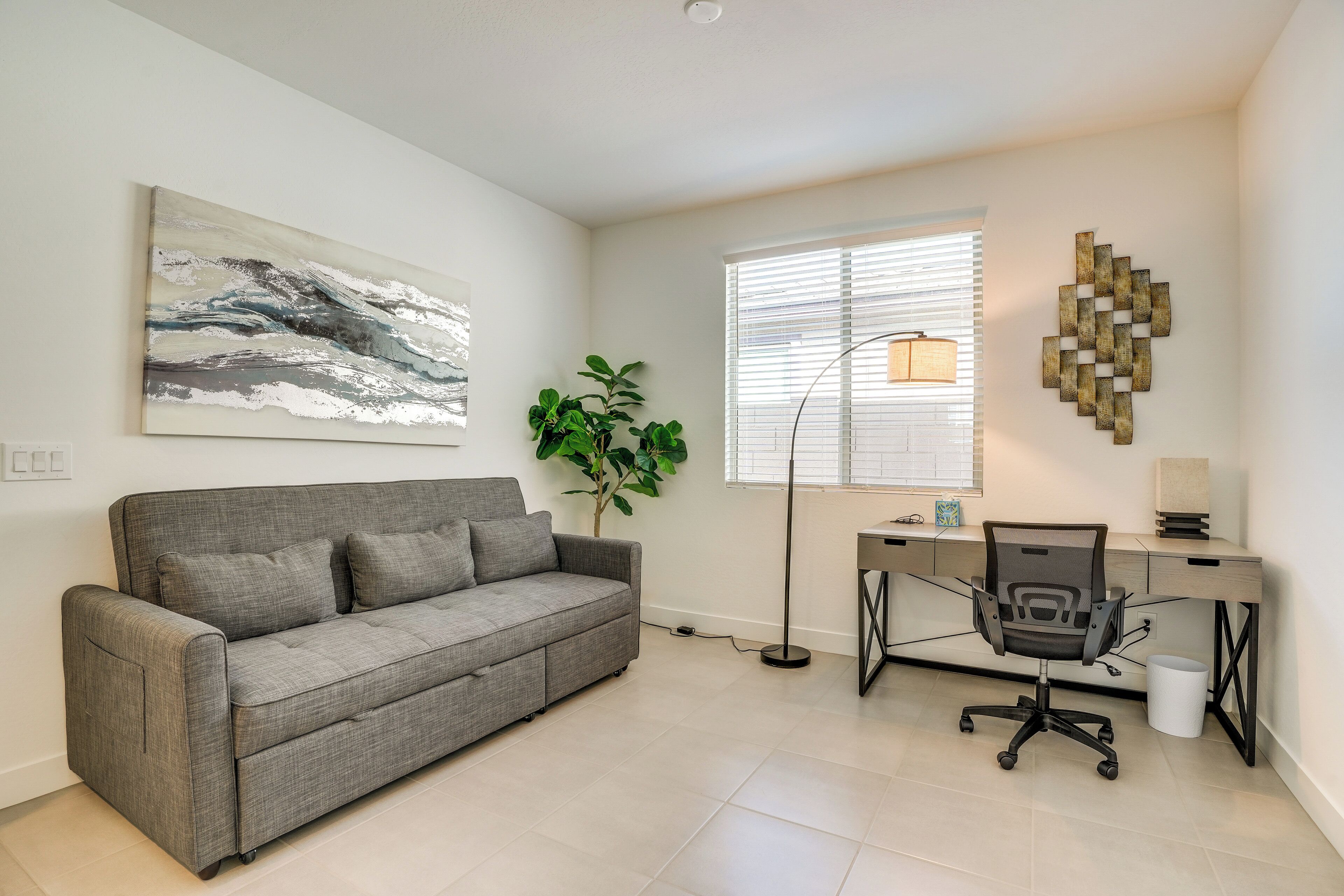 Un salon moderne avec un canapé gris, un bureau avec une chaise, une lampe sur pied et un grand tableau abstrait accroché au mur.