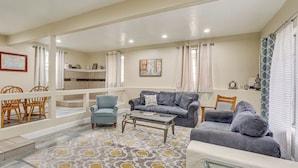 A spacious living area with a grey sofa, a blue armchair, and a glass coffee table. There is a dining area with wooden chairs and a table. The room has a patterned rug and a view of the kitchen through an open door. - Charming Lakehead Cabin: Walk to Shasta Lake! (Lakehead)